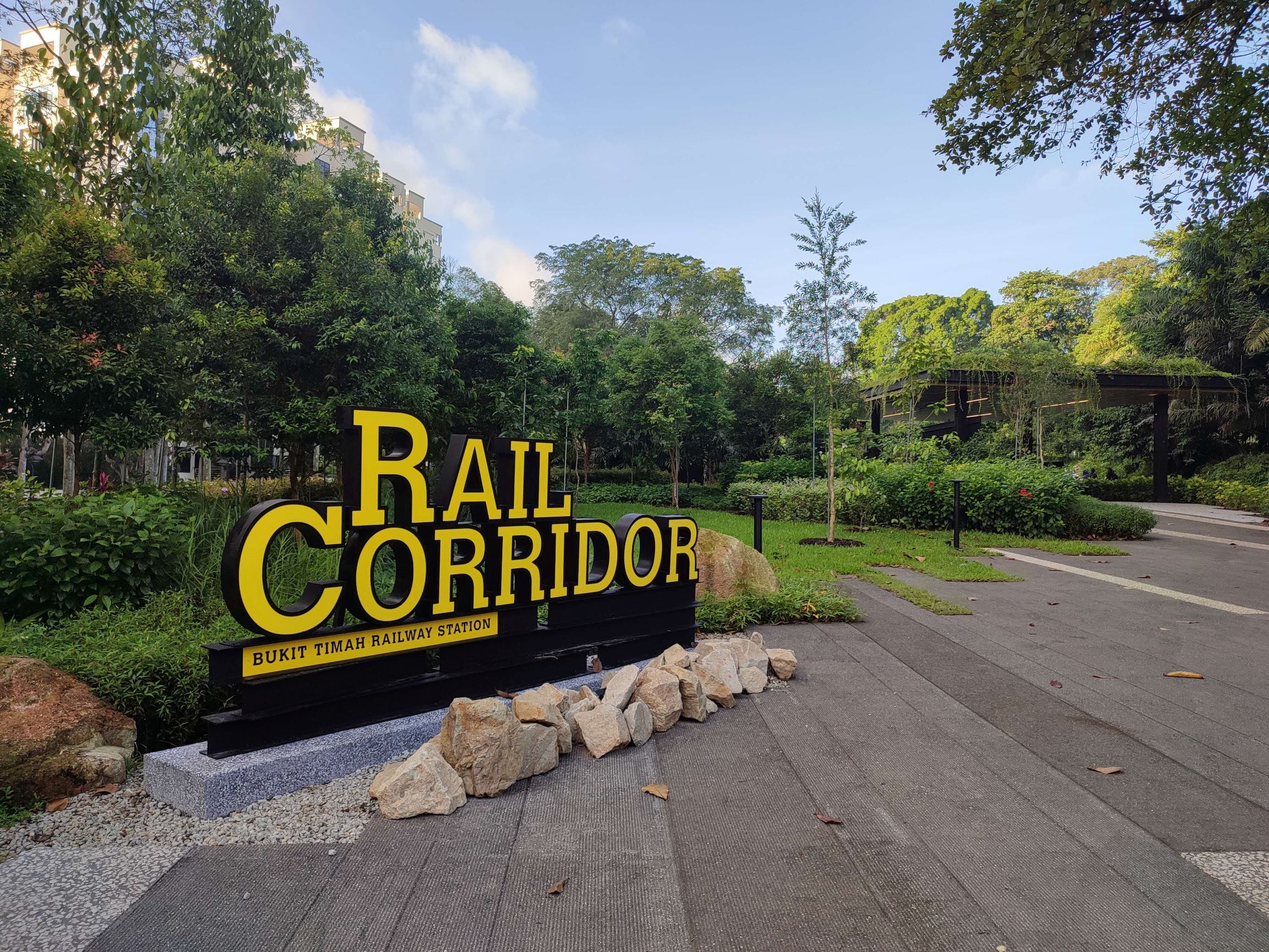 A yellow and black sign reads "Rail Corridor, Bukit Timah Railway Station" surrounded by trees and a paved pathway.